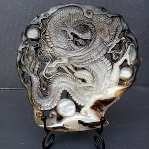 Può includere: Un intaglio circolare su conchiglia che presenta un intricato rilievo di drago con squame dettagliate, baffi fluenti e artigli. Il materiale iridescente naturale mostra sfumature di grigio, marrone e crema, accentuato da due sfere bianche lisce.