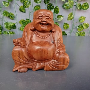May include: A wooden statue of a smiling Buddha sitting cross-legged with his hands in his lap. The statue is made of a light brown wood and has a natural finish.
