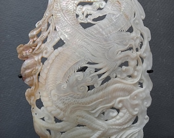 May include: A carved, pearlescent dragon sculpture. The dragon is intricately detailed, with scales, claws, and a flowing mane. The sculpture is mounted on a dark wooden stand, and the background is a neutral gray.