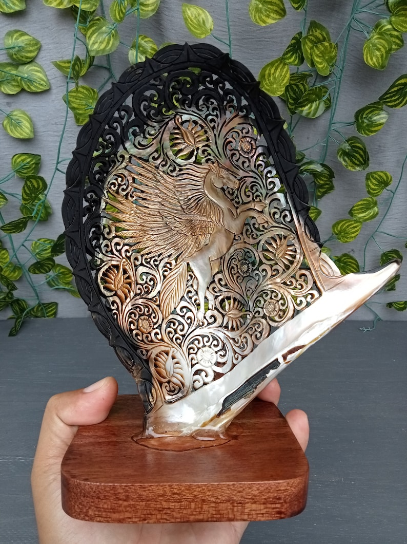 This is a real and natural mother of pearl shell polished and hand carved with wooden stand by a very skilled carving artist in his field. It is very detailed with pegasus design and has high artistic value