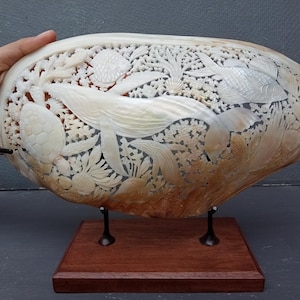 Könnte beinhalten: Eine große, oval geformte Muschel mit aufwendigen, handgeschnitzten Reliefdarstellungen von Meereslebewesen, darunter ein Wal, eine Meeresschildkröte und verschiedene Fische, die zwischen Korallenriffstrukturen schwimmen. Die Muschel steht auf einem Holzsockel.