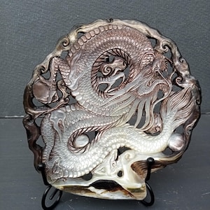 May include: A circular mother-of-pearl carving featuring an intricate dragon design with detailed scales, flowing whiskers, and a long tail. The shell displays iridescent shades of white, silver, and dark brown, with a polished finish and openwork.