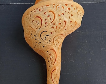 May include: A large, light orange conch shell featuring intricate, hand-carved filigree patterns across its surface. The shell has a spiraled top and a long, tapered body with delicate cut-out designs that reveal the hollow interior structure.