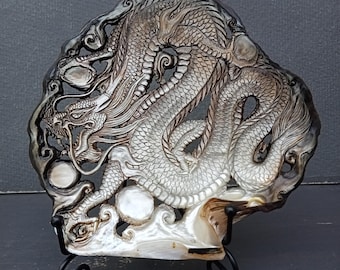 May include: A carved mother-of-pearl shell featuring an intricate dragon design with detailed scales, flowing whiskers, and swirling clouds. The iridescent surface displays shades of gray, white, and brown, supported by a black metal display stand.