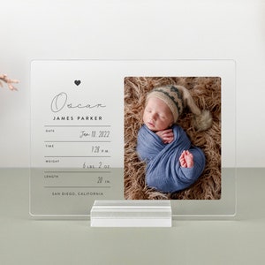 May include: A clear acrylic photo frame with a black heart and the text "Oscar James Parker" on the top. The frame includes a photo of a newborn baby sleeping in a blue blanket. The frame also includes the baby's birth date, time, weight, length, and location.