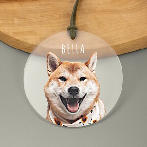 A round acrylic ornament showcasing a smiling dog in cartoon style. The name is written above in a white, playful font. The frosted ornament is attached with a green cord, adding a cheerful touch.