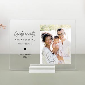 May include: A clear acrylic photo frame with a white background and black text that reads "Godparents are a blessing. Will you be mine? Love, Charlotte 2024". A black heart symbol is below the text. The frame has a photo of a family of three: a woman, a man, and a baby. The woman is wearing a white and black striped shirt. The man is wearing a white shirt. The baby is wearing a white and pink outfit. The family is smiling.