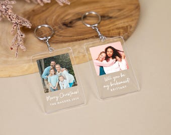 Könnte beinhalten: Zwei transparente Acryl-Foto-Schlüsselanhänger mit silbernen Schlüsselringen. Der erste Schlüsselanhänger hat ein Foto einer fünfköpfigen Familie und den Text "Frohe Weihnachten! Die Masons 2014". Der zweite Schlüsselanhänger hat ein Foto von zwei Frauen und den Text "Willst du meine Brautjungfer sein? Brittany".