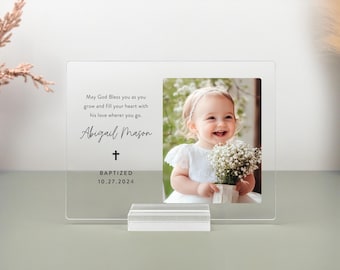 May include: A clear acrylic photo frame with a white background and black text. The text reads "May God Bless you as you grow and fill your heart with his love wherever you go. Abigail Mason + Baptized 10.27.2024". The frame has a photo of a young girl in a white dress holding a bouquet of white flowers.