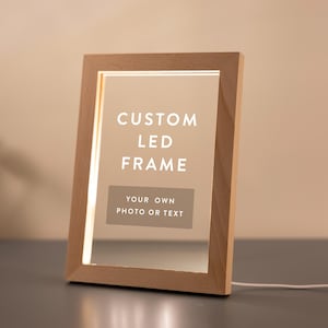Könnte beinhalten: Rechteckiger LED-Rahmen aus Holz mit dem Text "CUSTOM LED FRAME" und "YOUR OWN PHOTO OR TEXT" in Weiß. Der Rahmen ist von innen beleuchtet, und ein weißes Kabel erstreckt sich von der Basis. Eine kleine Topfpflanze ist im Hintergrund zu sehen.
