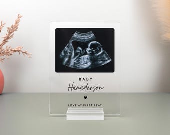 May include: Clear acrylic photo frame displaying a sonogram image of a baby. The frame includes the text "BABY Hanaderson" and "LOVE AT FIRST BEAT". The frame is supported by a clear acrylic base.