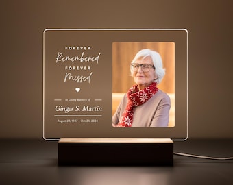 Könnte beinhalten: Eine weiße, beleuchtete Acryl-Gedenktafel mit einem Foto einer Frau mit Brille und rotem Schal. Der Text lautet "FOREVER Remembered FOREVER Missed" und "In Loving Memory of Ginger S. Martin August 24, 1947 - Oct 24, 2024".