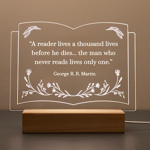 May include: A clear acrylic book-shaped night light with the quote "A reader lives a thousand lives before he dies... the man who never reads lives only one." by George R. R. Martin. The light sits on a wooden base and has floral accents.