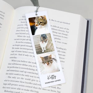 May include: A white bookmark featuring three photos of cats. The bookmark has a gray tassel and the words "Kitty Forever Loved" at the bottom. The bookmark is placed in an open book.