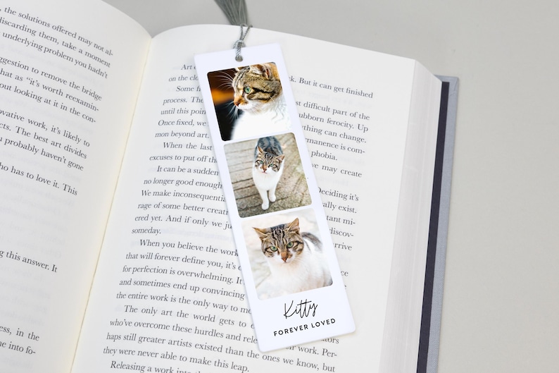 May include: A white bookmark featuring three photos of cats. The bookmark has a gray tassel and the words "Kitty Forever Loved" at the bottom. The bookmark is placed in an open book.