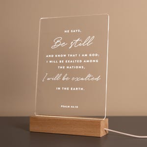 Personalized LED light featuring the calming Bible verse from Psalm 46:10: "He says, 'Be still and know that I am God; I will be exalted among the nations, I will be exalted in the earth.'" Engraved on a clear acrylic plaque with a warm wooden base, perfect for home décor, prayer spaces, or as a thoughtful Christian gift.