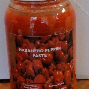 May include: A glass jar filled with red habanero pepper paste. The jar has a white lid and a label that reads "HABANERO PEPPER PASTE" with a picture of red peppers. The label also lists the ingredients: "Fresh Ground Peppers, Pickling Salt" and the farm information: "Zozoma Farms, Snellville, Georgia."
