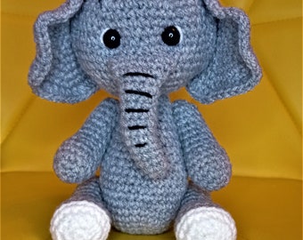 May include: A handmade gray crochet elephant toy with large ears, black eyes, and a trunk. The elephant has white feet and a small tuft of gray yarn on its head. The toy is sitting on a yellow surface.