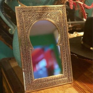 Puede incluir: Un espejo de metal dorado con un dise&ntilde;o ornamentado e intrincado. El espejo tiene una forma de arco tradicional marroqu&iacute; y est&aacute; enmarcado con un borde decorativo detallado.