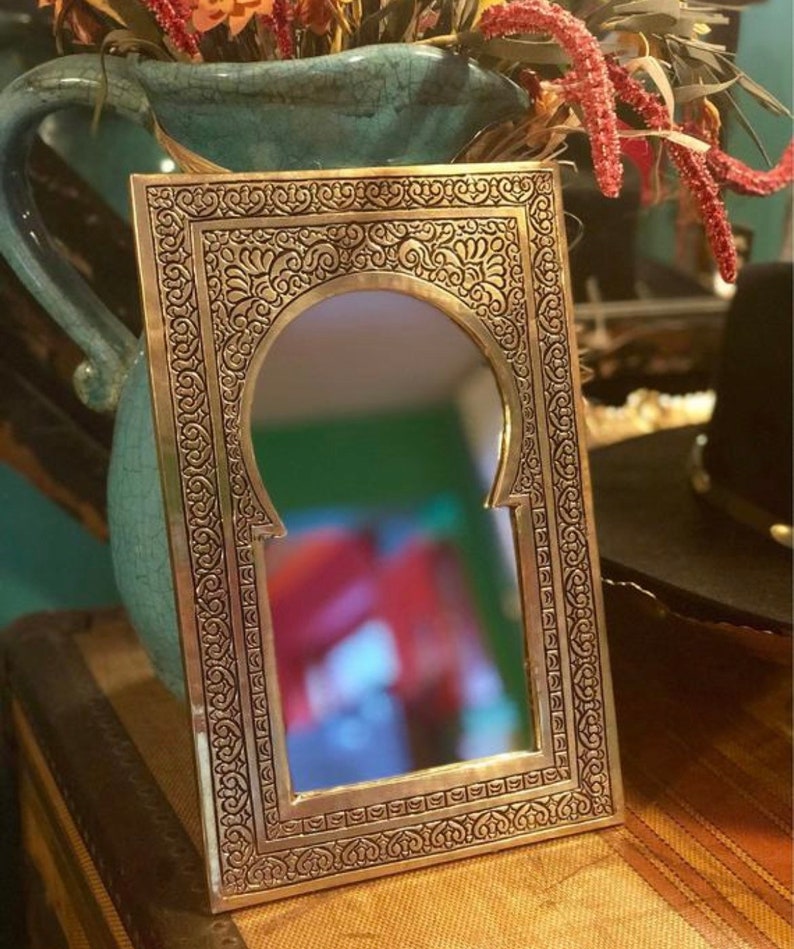 Puede incluir: Un espejo de metal dorado con un dise&ntilde;o ornamentado e intrincado. El espejo tiene una forma de arco tradicional marroqu&iacute; y est&aacute; enmarcado con un borde decorativo detallado.