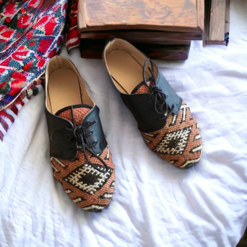 May include: A pair of handmade, patterned shoes with black leather accents. The shoes feature a woven design in shades of brown, beige, and black. The laces are black, and the shoes are set against a white background.
