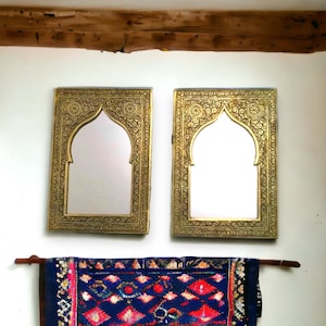May include: Two ornate gold-coloured framed mirrors with arched tops and intricate designs. A blue and red patterned rug hangs below the mirrors.