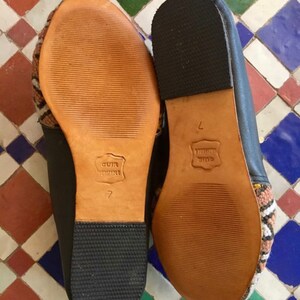 May include: Pair of black leather slippers with patterned fabric accents. The soles are light brown leather with the text "CUIR VERITABLE" and the number "7" embossed. The heels are black rubber.