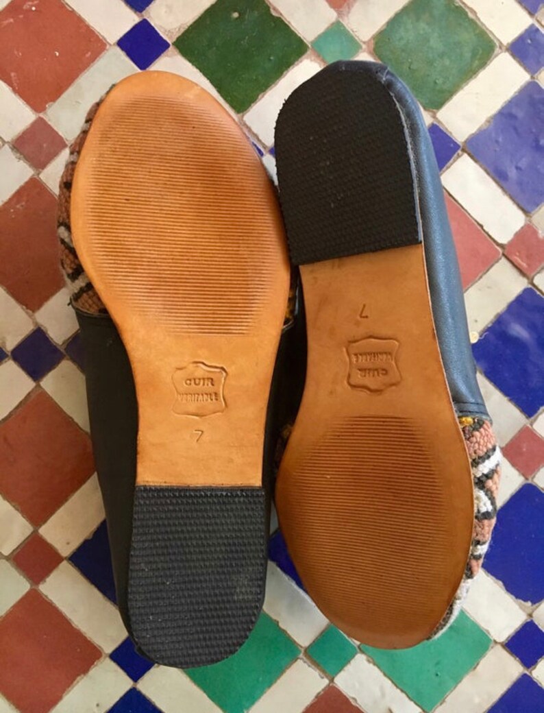 May include: Pair of black leather slippers with patterned fabric accents. The soles are light brown leather with the text "CUIR VERITABLE" and the number "7" embossed. The heels are black rubber.