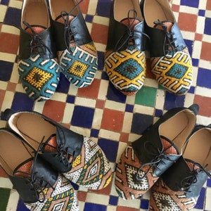 May include: A collection of six pairs of leather lace-up shoes. The shoes have black leather uppers and colorful, patterned fabric toe boxes. The patterns include geometric designs in shades of blue, yellow, brown, and white.