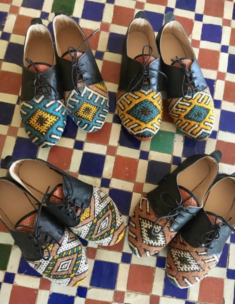 May include: A collection of six pairs of leather lace-up shoes. The shoes have black leather uppers and colorful, patterned fabric toe boxes. The patterns include geometric designs in shades of blue, yellow, brown, and white.