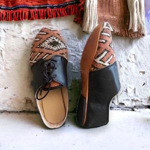 May include: Pair of handmade leather shoes. The shoes feature a brown and cream patterned fabric on the top, black leather accents, and laces. The soles are a light brown color. The shoes are displayed on a white surface.