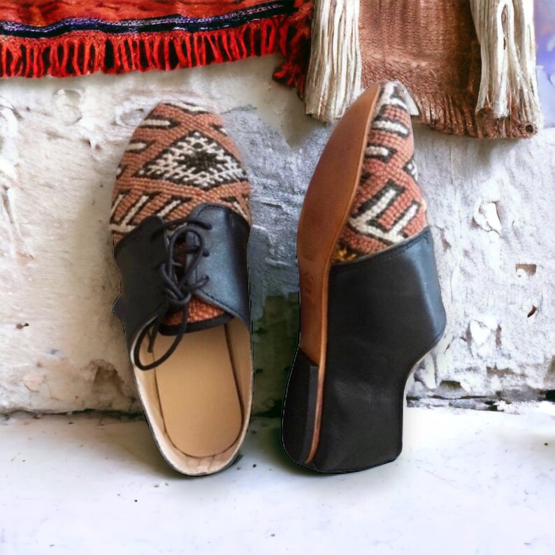 May include: Pair of handmade leather shoes. The shoes feature a brown and cream patterned fabric on the top, black leather accents, and laces. The soles are a light brown color. The shoes are displayed on a white surface.