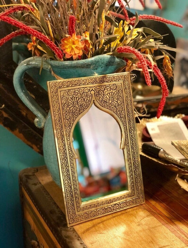 May include: A gold-toned metal mirror with an ornate, intricate design. The mirror has a rectangular frame with an arched top and a black background. The mirror is reflecting a blurry image of a room.