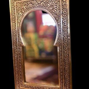 Puede incluir: Un espejo de metal dorado con un marco tallado ornamentado. El marco presenta un dise&ntilde;o de arco marroqu&iacute; tradicional con intrincados patrones florales.
