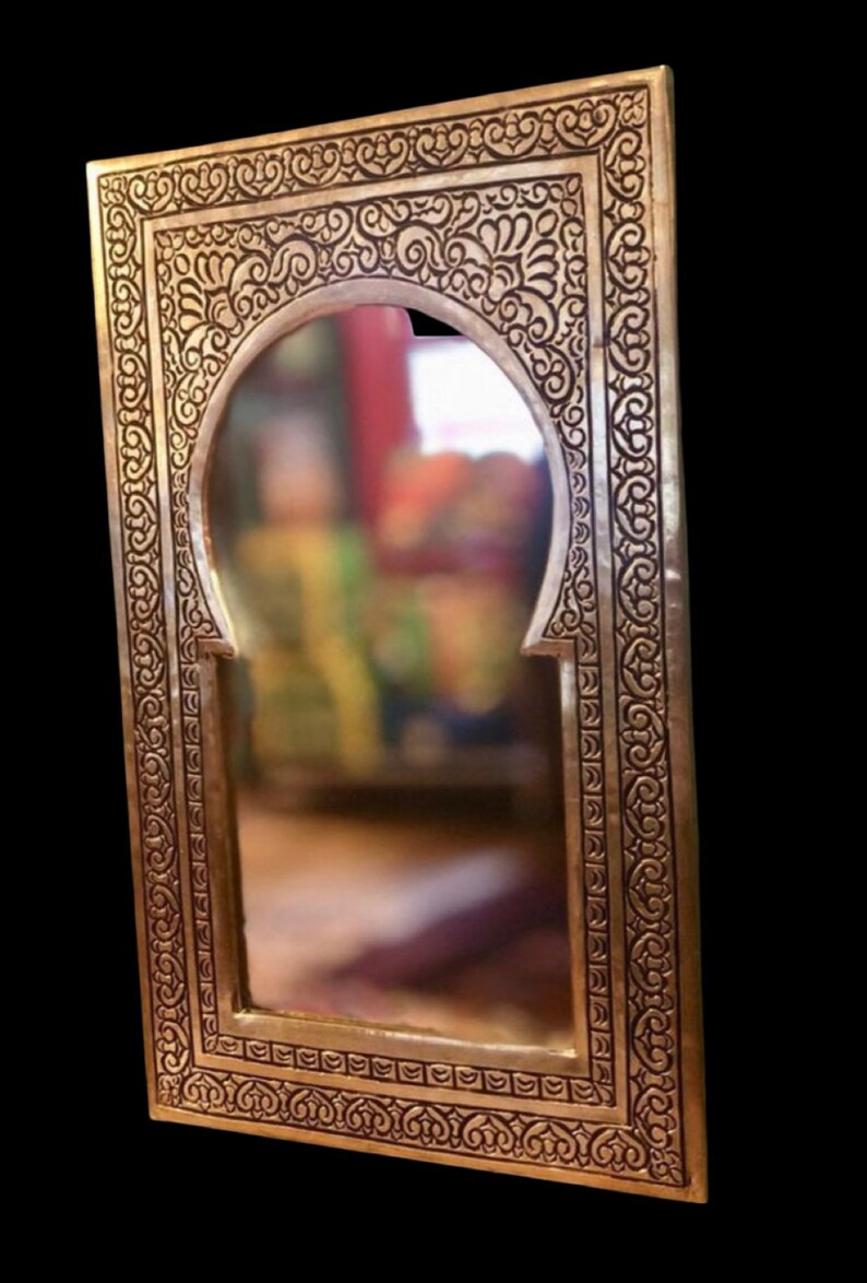Puede incluir: Un espejo de metal dorado con un marco tallado ornamentado. El marco presenta un dise&ntilde;o de arco marroqu&iacute; tradicional con intrincados patrones florales.
