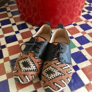 May include: A pair of handcrafted shoes with a unique design. The shoes feature black leather uppers and intricately woven fabric on the toe and sides, showcasing a geometric pattern in shades of orange, white, and black. The shoes are laced up with black laces.