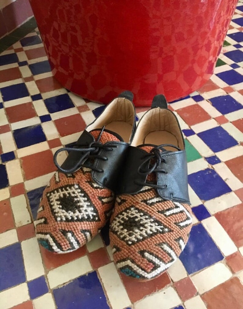 May include: A pair of handcrafted shoes with a unique design. The shoes feature black leather uppers and intricately woven fabric on the toe and sides, showcasing a geometric pattern in shades of orange, white, and black. The shoes are laced up with black laces.