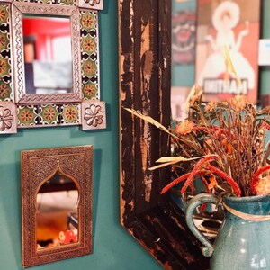 May include: Three decorative mirrors with ornate frames hang on a teal wall. The mirror on the left has a colourful tile design, the middle mirror has a brass frame with an arched top, and the mirror on the right has a dark wood frame with a distressed finish. A vase with dried flowers sits in front of the mirror on the right.