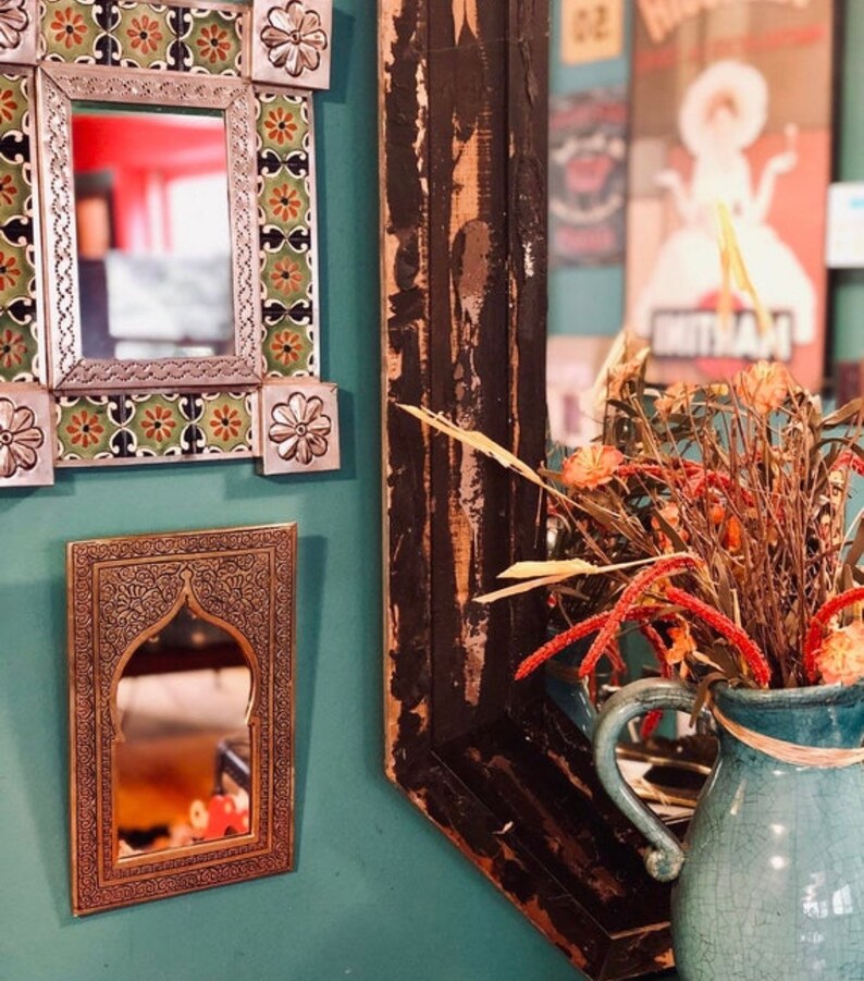 May include: Three decorative mirrors with ornate frames hang on a teal wall. The mirror on the left has a colourful tile design, the middle mirror has a brass frame with an arched top, and the mirror on the right has a dark wood frame with a distressed finish. A vase with dried flowers sits in front of the mirror on the right.