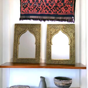 May include: Two ornate gold-coloured framed mirrors with arched tops, hanging on a white wall above a wooden shelf.