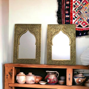 May include: Two ornate gold-coloured mirrors with arched tops and intricate carvings. The mirrors are displayed on a wooden shelf with decorative pottery.