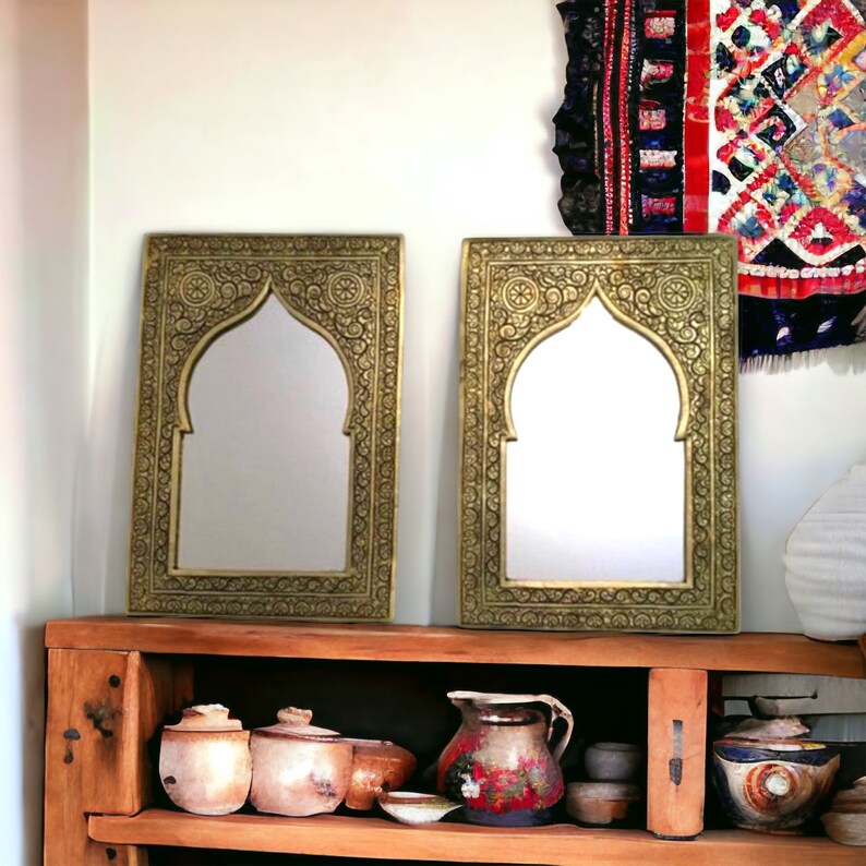 May include: Two ornate gold-coloured mirrors with arched tops and intricate carvings. The mirrors are displayed on a wooden shelf with decorative pottery.