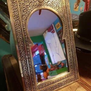 Puede incluir: Un espejo de metal dorado con un marco tallado ornamentado. El espejo tiene una parte superior arqueada y una superficie reflectante. El marco est&aacute; decorado con intrincados patrones florales.