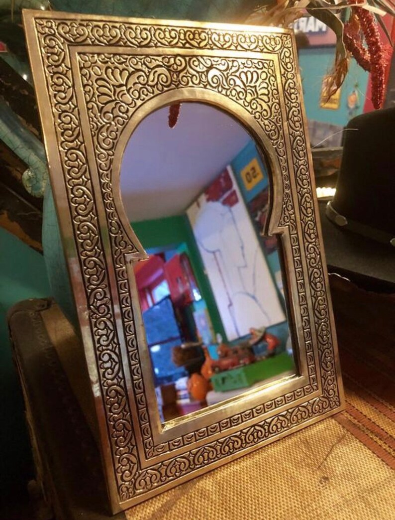 Puede incluir: Un espejo de metal dorado con un marco tallado ornamentado. El espejo tiene una parte superior arqueada y una superficie reflectante. El marco est&aacute; decorado con intrincados patrones florales.