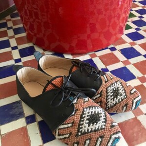 May include: A pair of black leather and woven fabric shoes with black laces. The woven fabric features a geometric pattern in shades of brown, white, and teal. The shoes are on a tiled floor with red, white, and blue tiles.