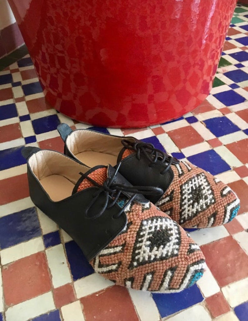 May include: A pair of black leather and woven fabric shoes with black laces. The woven fabric features a geometric pattern in shades of brown, white, and teal. The shoes are on a tiled floor with red, white, and blue tiles.