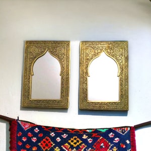 May include: Two ornate gold-coloured framed mirrors with arched openings. The mirrors are hanging on a white wall. A colourful patterned rug is hanging below the mirrors.