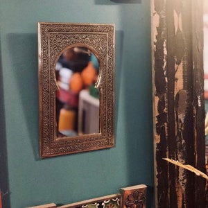 Puede incluir: Un espejo de metal plateado con un marco arqueado adornado. El espejo est&aacute; colgado en una pared azul verdosa con acentos de azulejos decorativos.