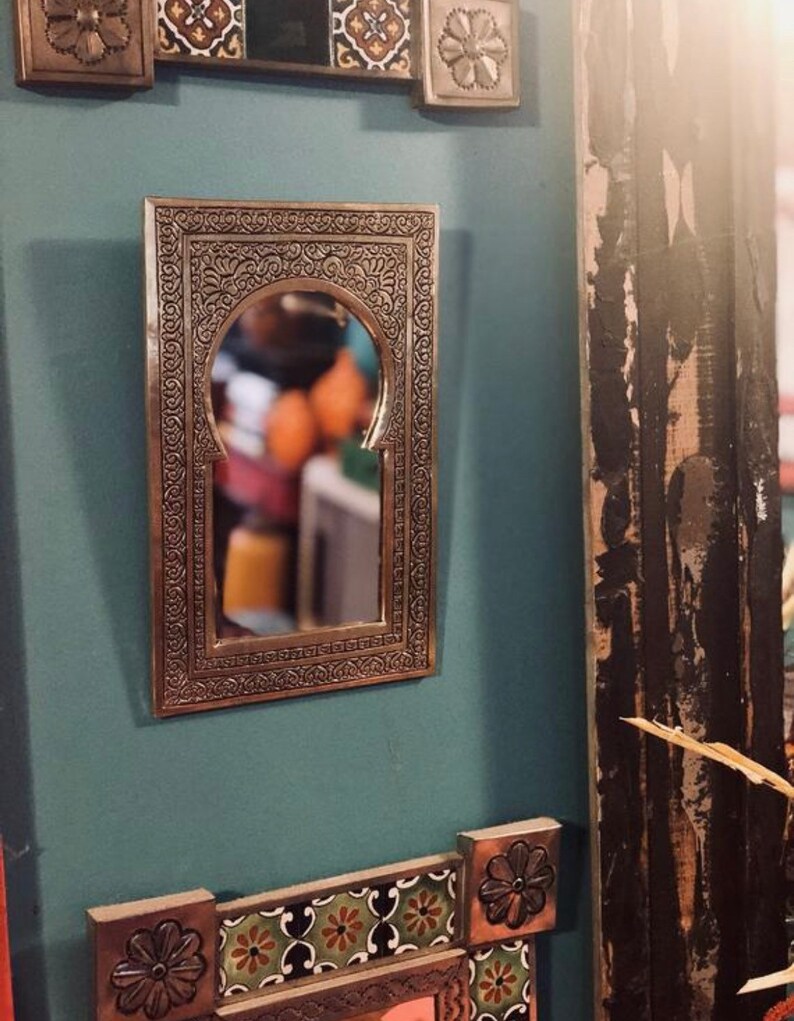 Puede incluir: Un espejo de metal plateado con un marco arqueado adornado. El espejo est&aacute; colgado en una pared azul verdosa con acentos de azulejos decorativos.