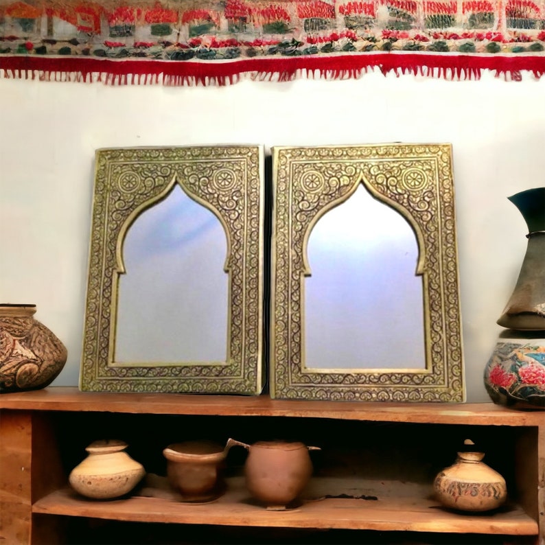 May include: Two ornate gold-coloured mirrors with intricate carvings and arched tops. The mirrors are set against a white wall and a wooden shelf with various decorative items.